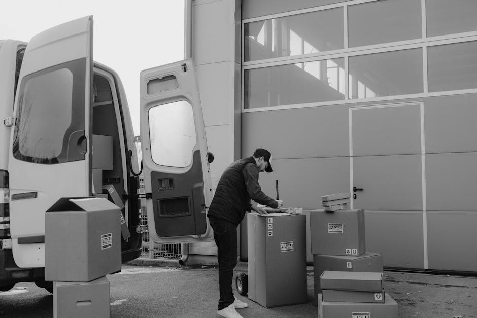 A man wearing a cap and dark clothing is standing outside near a large commercial van with its rear doors open, engaged in packing or preparing items for a home relocation. In front of him are several cardboard boxes, some sealed with tape and others stacked, along with a few small packages and documents. The boxes are positioned on the pavement next to the van, which is parked on a street adjacent to a modern building with large glass windows and metal panels. The man is handling papers or labels on a box, possibly organizing or verifying contents as part of the packing process. Inside the van, visible through the open rear doors, there are additional packing materials and possibly furniture or equipment for furniture transport, indicative of a professional removal service. Man With a Van Aldersbrook, a removals company, appears to be handling loading or packing tasks at this site, supporting efficient moving logistics and household packing and moving practices.