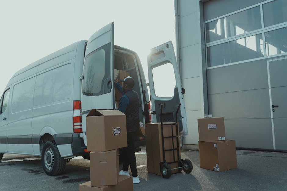 A professional mover from Man With a Van Aldersbrook is seen loading cardboard boxes and packing materials into the rear of a white panel van outside a modern building with large glass windows. The boxes are of various sizes, some sealed with packing tape and marked with labels, while others are open revealing packed contents inside. The mover is wearing a dark uniform and a cap, using a hand cart to assist with transporting the boxes. The loading process takes place in a designated loading bay area with a paved surface and ample space for moving furniture and household items during a home relocation. The background includes the building's exterior and sunlight illuminating the scene, emphasizing the organized logistics involved in furniture transport and packing as part of professional removals services offered by Man With a Van Aldersbrook.