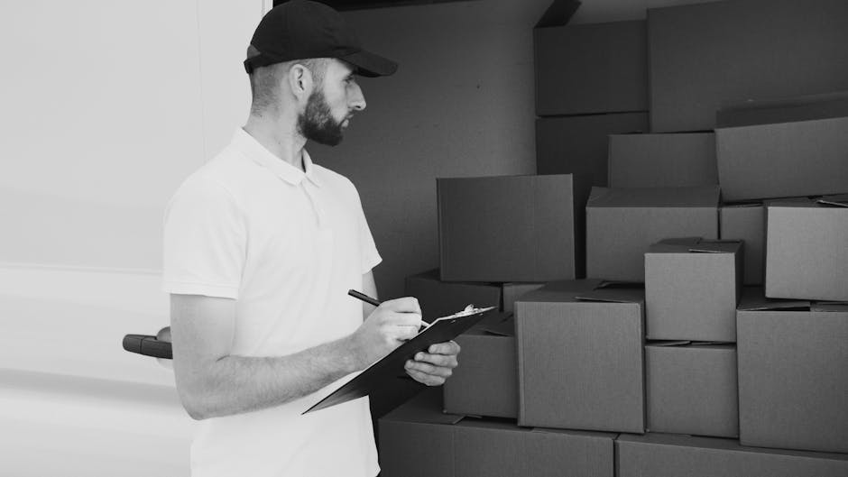 Two men preparing for home relocation by loading a large cardboard box into the back of a white van parked outside a residential property. One man is wearing a black padded vest over a blue long-sleev
