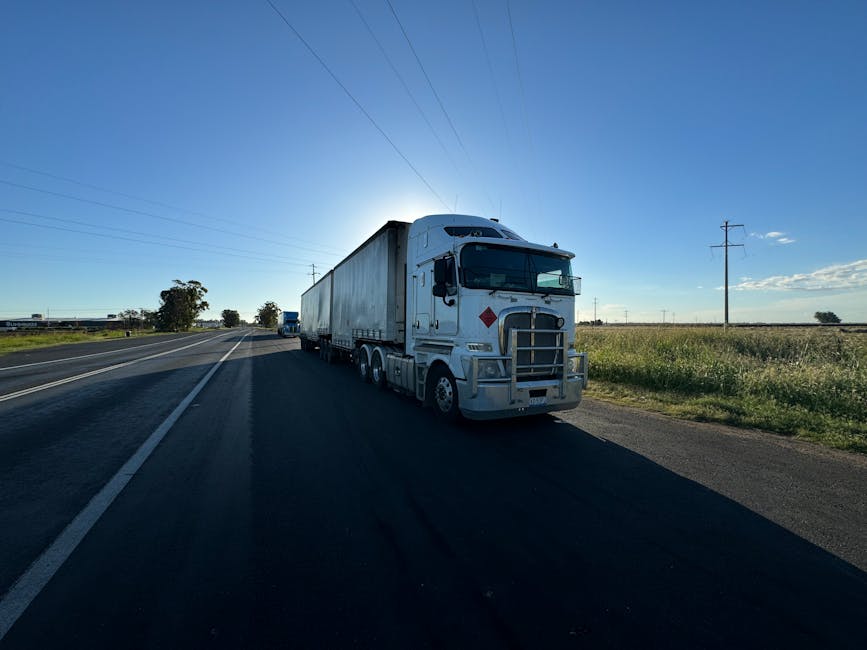 A large white commercial freight truck with a spacious trailer is parked on the side of a rural road during daylight hours, with the sun low in the sky creating a bright glare behind the vehicle. The truck is positioned partially on the paved shoulder, close to a grassy verge, with a clear sky and a few scattered clouds overhead. Utility poles and power lines run parallel to the road, extending into the distance on both sides. The trailer appears empty or used for transporting furniture and household items during a house removal, as the scene suggests a relocation process. No other vehicles or people are visible in the immediate vicinity, emphasizing the truck's role in home relocation logistics. The environment indicates a quiet, open area suitable for loading or unloading furniture and packing materials, supported by the professional services offered by Man With a Van Aldersbrook for removals and moving logistics.