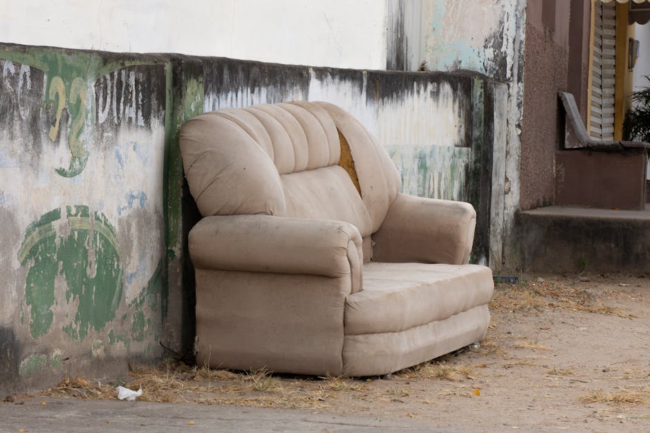 A worn beige armchair with visible fabric wear and a torn section on the backrest is positioned outside on a dirt and gravel surface, leaning against an aged concrete wall with faded green and black paint and peeling patches. The surrounding area includes a set of concrete stairs leading to a door on the right side of the image, and a small potted plant is partially visible near the entrance. The scene appears to be part of a household move or item disposal process, with the armchair likely awaiting collection or transport by Man With a Van Aldersbrook as part of home relocation or furniture transport services. The lighting is natural, highlighting the texture of the fabric and the weathered condition of the furniture and the environment, consistent with an outdoor home clearance or packing and moving setup.