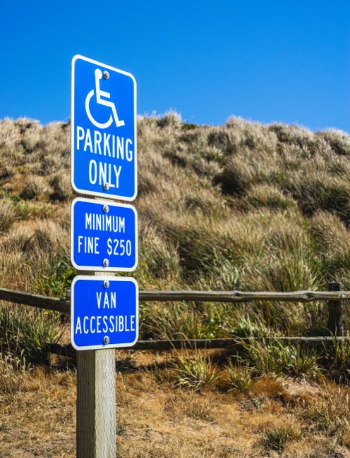 A close-up view of a blue parking sign in an outdoor setting, featuring a white wheelchair symbol at the top indicating designated parking for disabled persons. Below, additional blue signs display the words 'PARKING ONLY,' 'MINIMUM FINE £250,' and 'VAN ACCESSIBLE.' The signpost is mounted on a metal pole situated on a paved area, with dry grass, sand dunes, and wooden fencing in the background under a clear blue sky, suggesting a coastal or natural environment. The signage is clearly visible, serving as a visual marker for accessible parking and vehicular regulations, relevant to vehicle access and loading considerations in house removals and relocation logistics.
