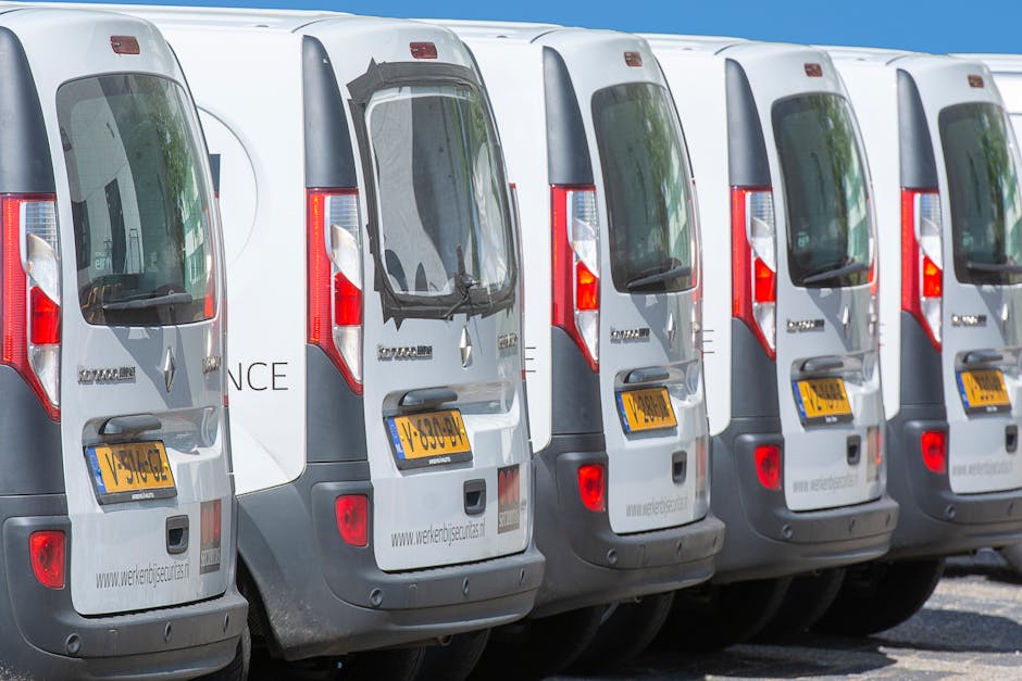 A row of five white vans with tinted rear windows and yellow registration plates parked side by side on a paved surface under a clear blue sky. The vans belong to Man With a Van Aldersbrook, a UK moving and removals company, and are aligned to facilitate home relocation and furniture transport services. The rear doors of each van are closed, with some displaying branding and website information. The image captures the end view of the vehicles, showing details such as tail lights, handles, and reflective surfaces. The setting appears to be a designated loading area or parking space used for loading household items, boxes, and furniture during a packing and moving process. The environment indicates a professional and organized vehicle fleet ready for household removals and relocation logistics.