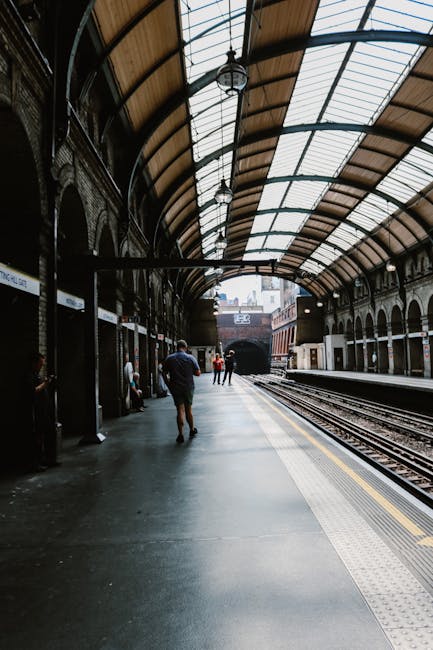 Inside a historic train station with a curved glass and metal roof structure, featuring a platform with train tracks running alongside. Several passengers are visible, some standing and waiting, while others walk along the platform. The platform surface is made of smooth concrete with tactile paving near the edge for safety. On the left, there are small shelters with benches, and a few people are seated. The lighting is primarily natural, filtering through the glass roof, creating a bright, well-lit environment suitable for a home relocation or furniture transport process. The station setting underscores the importance of efficient moving logistics, as outlined in the Forest Gate Station moves page of Man With a Van Aldersbrook, who provides professional removals and packing services for house moves, ensuring smooth transportation of belongings through such transit hubs.