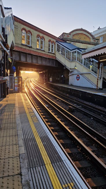 No visible furniture, boxes, or packaging materials related to house moves are present in this image. The photo captures a railway station platform at sunset, with a yellow tactile paving strip along the edge and a set of train tracks running parallel. An overhead bridge with staircases and enclosed walkways crosses above the tracks, leading to the station entrance. The station structure includes brick and concrete elements, with a small London Underground logo visible on the side of the bridge. The environment appears clean and well-lit, with natural sunlight casting a warm glow on the tracks and nearby buildings. This setting relates to the logistical aspects of home relocation or furniture transport processes, as it depicts a transportation hub where luggage or items could be transferred or prepared for transport, aligning with the services of companies like Man With a Van Aldersbrook.