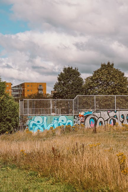 A metal fence with vertical bars and a graffiti-covered panel separates a grassy area with tall, dry grass and wildflowers from an outdoor space. Behind the fence, there are several large, leafy trees and a modern brick residential building with balconies. The sky above is partly cloudy with large, white clouds and patches of blue. This scene relates to home relocation and furniture transport processes, as it reflects the type of environment where a professional removals service like Man With a Van Aldersbrook might operate during a house move near Wanstead Flats, involving loading and unloading furniture and boxes across outdoor spaces and urban surroundings.