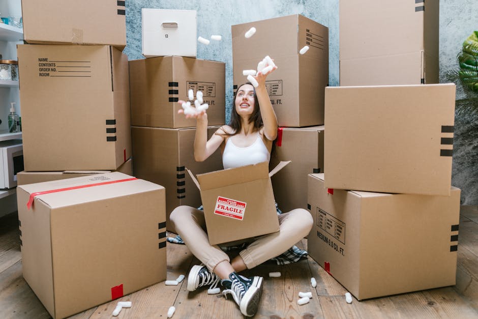 A young woman with long dark hair, wearing a white sleeveless top, beige pants, and black sneakers, is sitting cross-legged on a wooden floor inside a room, surrounded by large cardboard boxes for relocation. She is smiling and holding handfuls of white packing foam dunnage, which are falling into an open box in her lap. The boxes vary in size, some sealed with red packing tape and others stacked against the blue and grey textured walls of the room. A small green plant is visible in the corner, and a white shelf with household items is partially seen on the left. The scene depicts the packing stage of a home relocation, with the foam packing material used for furniture and fragile items protection, reflecting a professional removals process by [COMPANY_NAME]. The lighting is bright and natural, illuminating the area for efficient packing and moving activities typical of same-day Aldersbrook removals for urgent relocations, as described on the webpage.