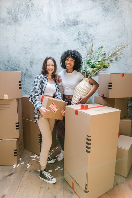 A young woman with long dark hair, wearing a white sleeveless top, beige pants, and black sneakers, is sitting cross-legged on a wooden floor inside a room, surrounded by large cardboard boxes for relocation. She is smiling and holding handfuls of white packing foam dunnage, which are falling into an open box in her lap. The boxes vary in size, some sealed with red packing tape and others stacked against the blue and grey textured walls of the room. A small green plant is visible in the corner, and a white shelf with household items is partially seen on the left. The scene depicts the packing stage of a home relocation, with the foam packing material used for furniture and fragile items protection, reflecting a professional removals process by [COMPANY_NAME]. The lighting is bright and natural, illuminating the area for efficient packing and moving activities typical of same-day Aldersbrook removals for urgent relocations, as described on the webpage.
