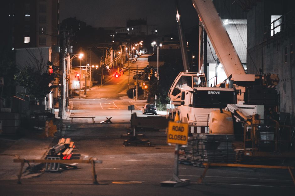 A nighttime view of a street closure area for home relocation in Aldersbrook, showing a construction site with a large white crane parked on the right side of the road. The crane is positioned next to a building with a partially visible doorway. In the foreground, there are several orange and yellow 'Road Closed' signs, along with barriers and traffic cones blocking the street. The street is illuminated by several streetlights, casting warm light on parked cars and the surrounding residential buildings further uphill. Some small stacks of wooden pallets and metal barriers are placed near the crane, indicating ongoing loading and unloading activities related to furniture transport and packing for a move. The scene captures an active moving logistics environment, managed by Man With a Van Aldersbrook, with visible elements supporting efficient execution of house removals and relocation services.