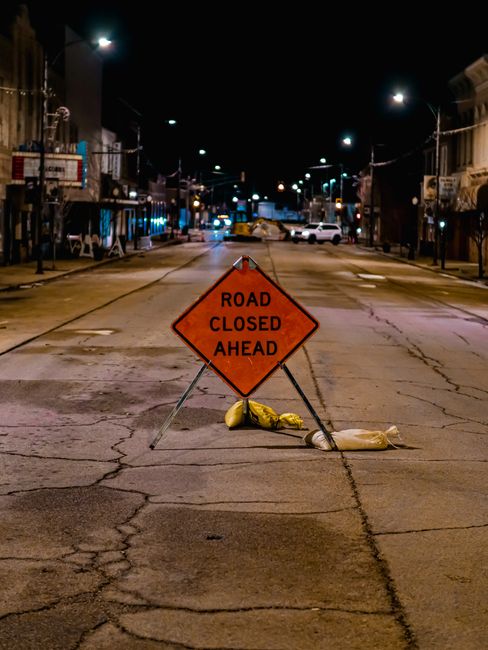 A nighttime street scene showing a temporary orange road closure sign with the message 'ROAD CLOSED AHEAD' positioned in the middle of an asphalt road. The sign is supported by metal legs and is accompanied by yellow and beige sandbags to keep it in place. The street is illuminated by overhead streetlights, with visible cracks and patches on the pavement. On either side of the road, there are residential buildings with facades and balconies, some displaying shop fronts or entrances. Parked cars line both sides of the street, and the road appears to be clear of traffic. The setting suggests a residential area undergoing a home relocation or moving process, potentially related to the street closure regulations for Aldersbrook moves, as managed by Man With a Van Aldersbrook, with the scene captured as part of the logistics involved in packing, loading, and transport during a removal service.