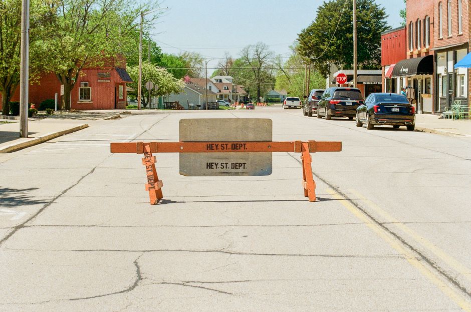A nighttime view of a street closure area for home relocation in Aldersbrook, showing a construction site with a large white crane parked on the right side of the road. The crane is positioned next to a building with a partially visible doorway. In the foreground, there are several orange and yellow 'Road Closed' signs, along with barriers and traffic cones blocking the street. The street is illuminated by several streetlights, casting warm light on parked cars and the surrounding residential buildings further uphill. Some small stacks of wooden pallets and metal barriers are placed near the crane, indicating ongoing loading and unloading activities related to furniture transport and packing for a move. The scene captures an active moving logistics environment, managed by Man With a Van Aldersbrook, with visible elements supporting efficient execution of house removals and relocation services.