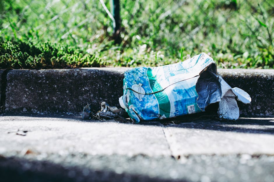 A crumpled piece of aluminum foil and a torn plastic bag containing various waste items are placed on a concrete curb on the pavement, with green grass and plants in the background. The scene is outdoors in natural daylight, and the waste appears discarded deliberately or accidentally, reflecting the need for rubbish collection services. The plastic bag is partially open, revealing some debris inside, and the foil wrapper shows signs of damage. This image, associated with house removals and relocation services by Man With a Van Aldersbrook, emphasizes proper waste disposal during home moving processes and the importance of organized rubbish clearance in Aldersbrook, especially when preparing for a home relocation or furniture transport involving loading or unloading outdoor areas.