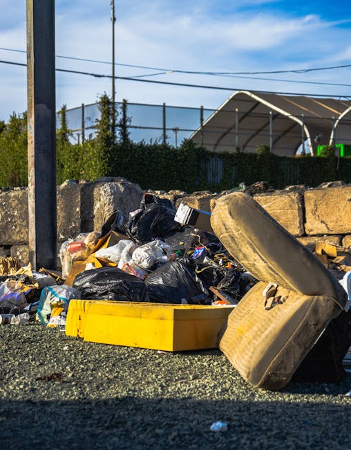 A crumpled piece of aluminum foil and a torn plastic bag containing various waste items are placed on a concrete curb on the pavement, with green grass and plants in the background. The scene is outdoors in natural daylight, and the waste appears discarded deliberately or accidentally, reflecting the need for rubbish collection services. The plastic bag is partially open, revealing some debris inside, and the foil wrapper shows signs of damage. This image, associated with house removals and relocation services by Man With a Van Aldersbrook, emphasizes proper waste disposal during home moving processes and the importance of organized rubbish clearance in Aldersbrook, especially when preparing for a home relocation or furniture transport involving loading or unloading outdoor areas.